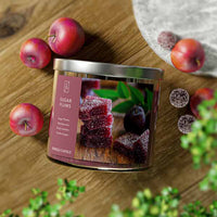 A three-wick candle in a clear glass jar with a silver lid and a festive holiday-themed label. The label features a bowl of sugared plums and juicy blackberries, dusted with powdered sugar. The candle’s wax is deep purple, and the wicks are unlit. The scent notes listed are sugared plums, blackberries, delicate florals, amber, and vanilla sugar.