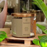 A three-wick candle in a clear glass jar with a black lid and a sophisticated wood-themed label. The label features a polished teakwood surface with an elegant woodgrain pattern. The candle’s wax is rich brown, and the wicks are unlit. The scent notes listed are burlwood, cedarwood, oak, musk, and rosemary.