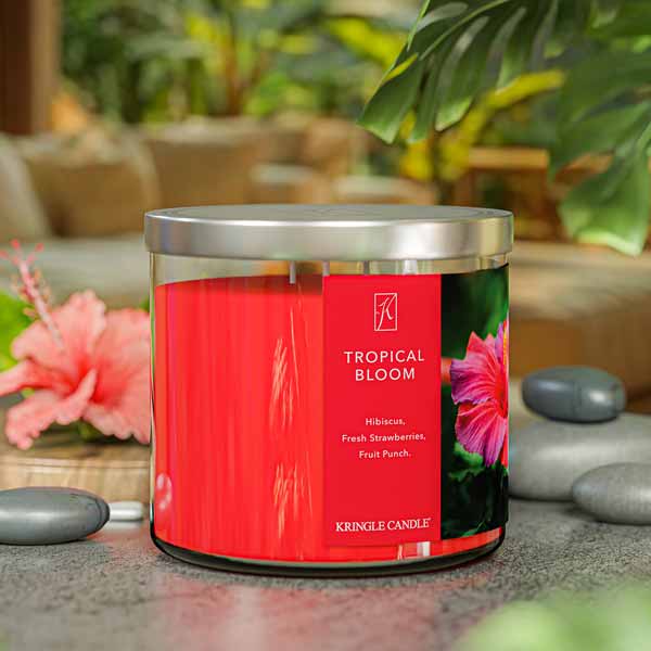 A three-wick candle in a clear glass jar with a gold lid and a vibrant floral-themed label. The label features a lush tropical arrangement of hibiscus flowers, bright pink and red petals, and green leaves. The candle’s wax is hot pink, and the wicks are unlit. The scent notes listed are hibiscus, strawberries, and fruit punch.