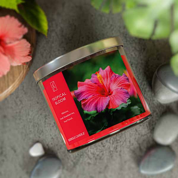 A three-wick candle in a clear glass jar with a gold lid and a vibrant floral-themed label. The label features a lush tropical arrangement of hibiscus flowers, bright pink and red petals, and green leaves. The candle’s wax is hot pink, and the wicks are unlit. The scent notes listed are hibiscus, strawberries, and fruit punch.