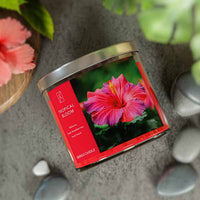 A three-wick candle in a clear glass jar with a gold lid and a vibrant floral-themed label. The label features a lush tropical arrangement of hibiscus flowers, bright pink and red petals, and green leaves. The candle’s wax is hot pink, and the wicks are unlit. The scent notes listed are hibiscus, strawberries, and fruit punch.