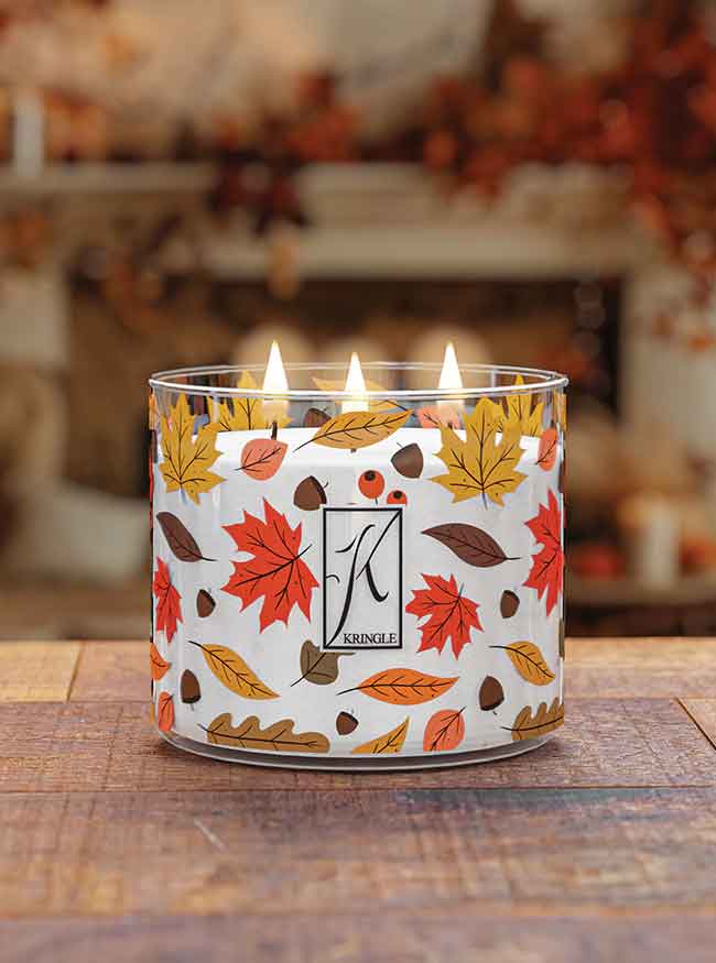 A lit candle in a glass jar with autumn leaves design on a wooden table, blurred autumnal decor in the background.