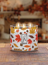 A lit candle in a glass jar with autumn leaves design on a wooden table, blurred autumnal decor in the background.