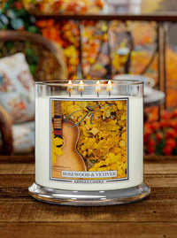 Lit 4-wick candle in a clear jar with a label of an acoustic guitar amidst yellow autumn leaves.