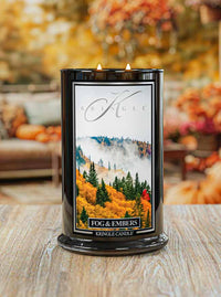Kringle Candle with 'Fog & Embers' packaging on a wooden surface with blurred autumn decorations in the background.
