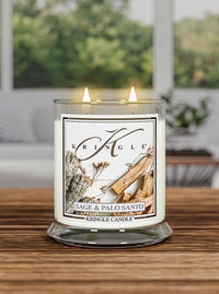 Lit Kringle candle labeled "Sage & Palo Santo" on a wooden table with a blurred green outdoor background.