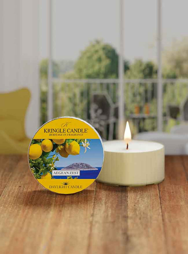 Lit white candle beside a "Kringle Candle" tin with a lemon design, labeled "Aegean Zest," on a wooden table.