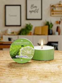 Green candle tin labeled "Fresh Bergamot" next to a lit candle on a wooden table; blurred kitchen background.