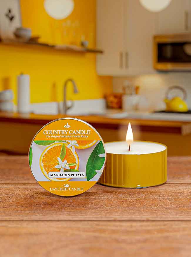 A lit candle labeled "Country Candle, Mandarin Petals" on a wooden surface in a bright, yellow-themed kitchen.
