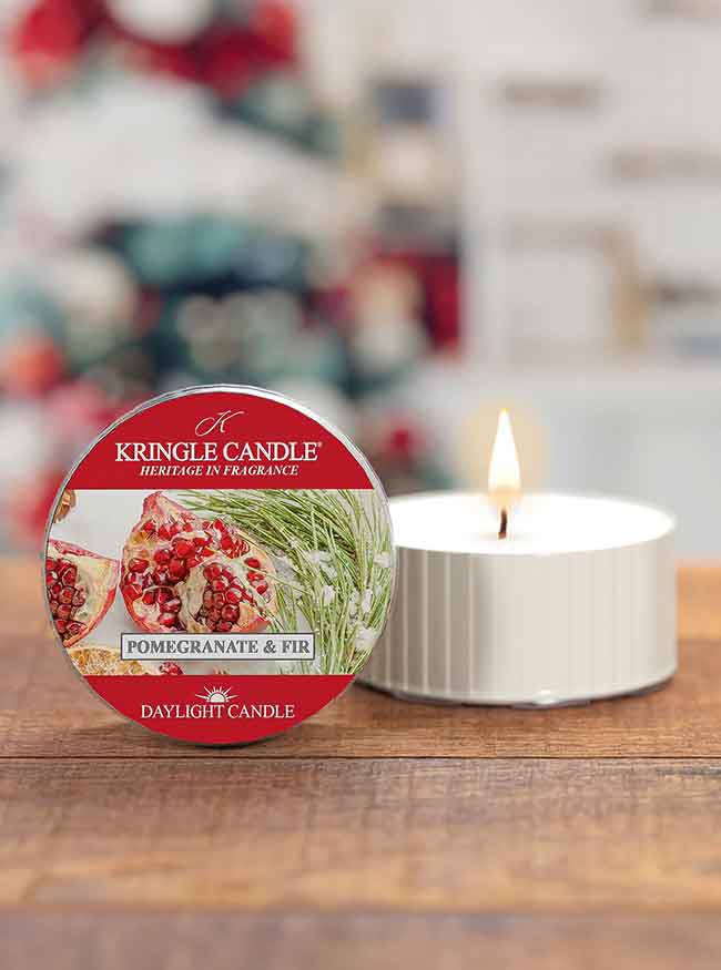 Red tin for "Pomegranate & Fir" candle, next to a lit white candle on a wooden surface with a blurred festive background.