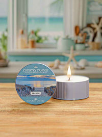 Lit white candle in a tin labeled "Country Candle Seaside" on a wooden table with blurred beach-themed background.