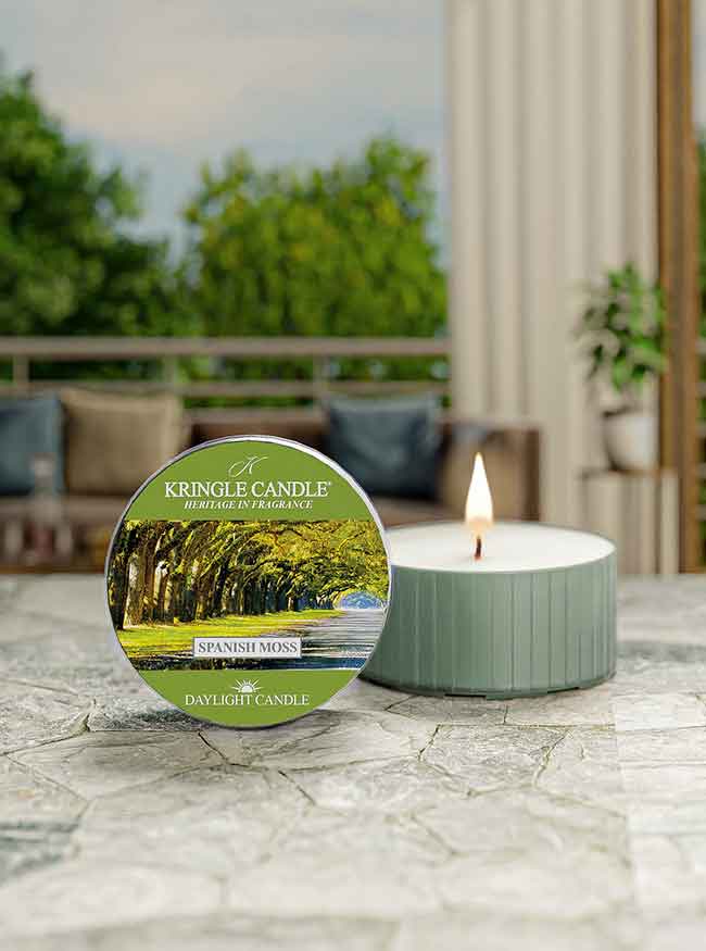 A lit tealight candle labeled "Kringle Candle Spanish Moss" on a stone surface, with a blurred outdoor background.