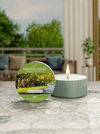 A lit tealight candle labeled "Kringle Candle Spanish Moss" on a stone surface, with a blurred outdoor background.