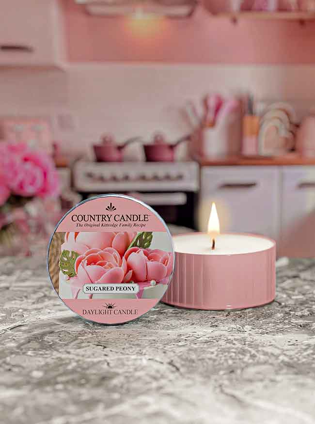 Pink scented candle labeled "Sugared Peony" on marble surface, with a cozy kitchen background.