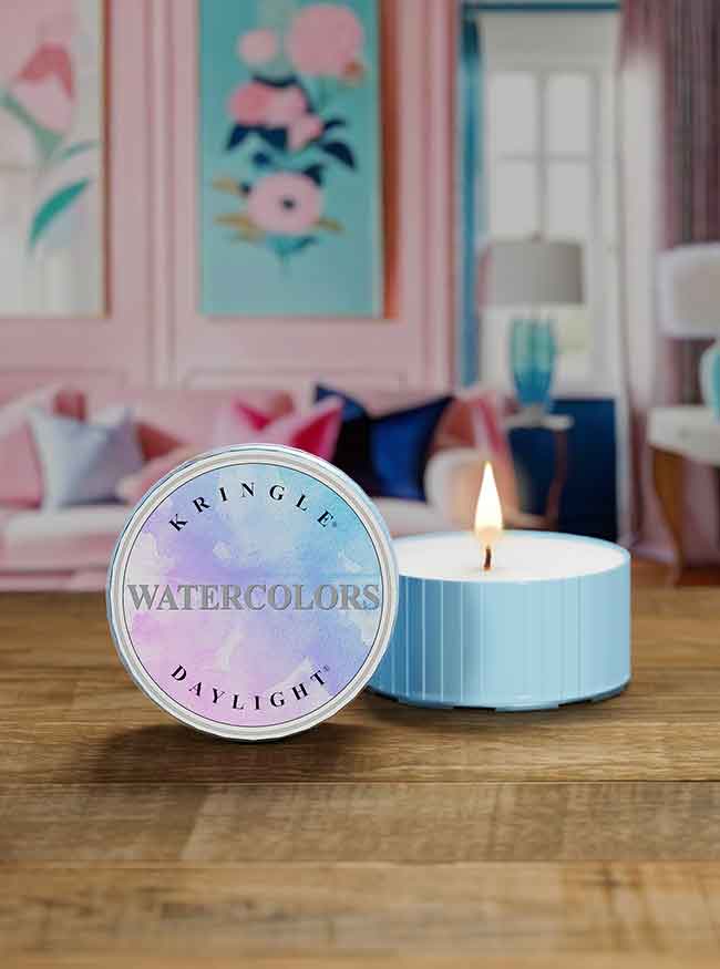 A Kringle Candle Daylight in the “Watercolors” scent displayed on a wooden surface. The candle has a light blue ribbed container, with one unit lit and another showing the lid featuring a soft pastel watercolor design. In the background, there’s a cozy living space with pink and blue decor, floral wall art, and plush cushions, creating a soft, artistic ambiance.