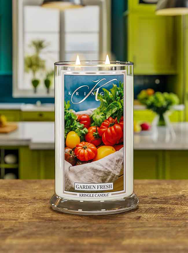 A lit candle labeled "Garden Fresh" with images of tomatoes and greens, set on a wooden table in a kitchen.