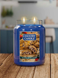 Blue candle labeled "Country Candle, Glazed Berry Scone" with two lit wicks, placed on a wooden table.