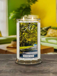 A lit candle labeled "Spanish Moss" by Kringle Candle, with a scenic forest image, surrounded by a living room background.