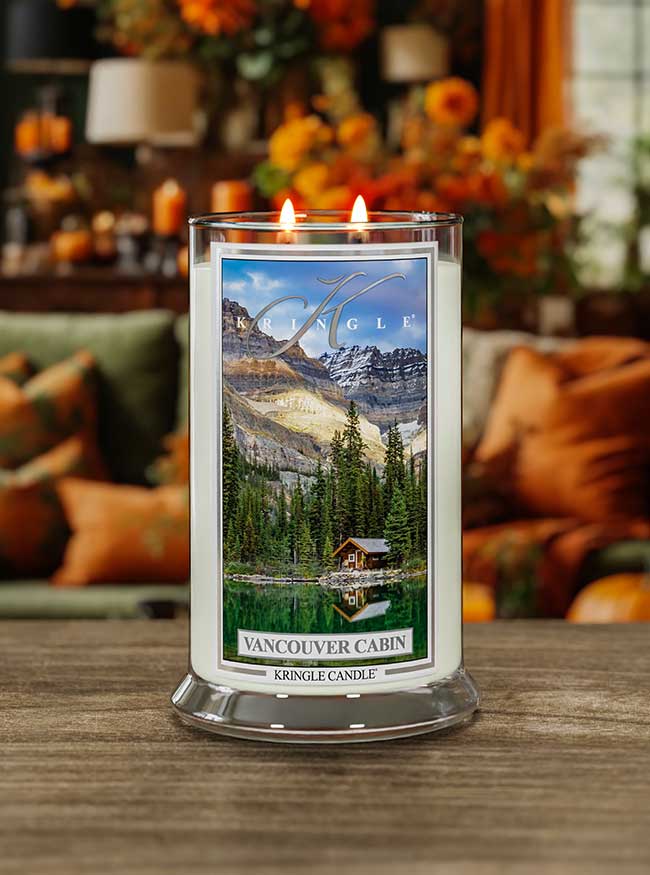 Lit Vancouver Cabin 2-wick candle. Label shows a cabin by a forest and lake with mountains in the background.