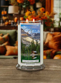 Lit Vancouver Cabin 2-wick candle. Label shows a cabin by a forest and lake with mountains in the background.