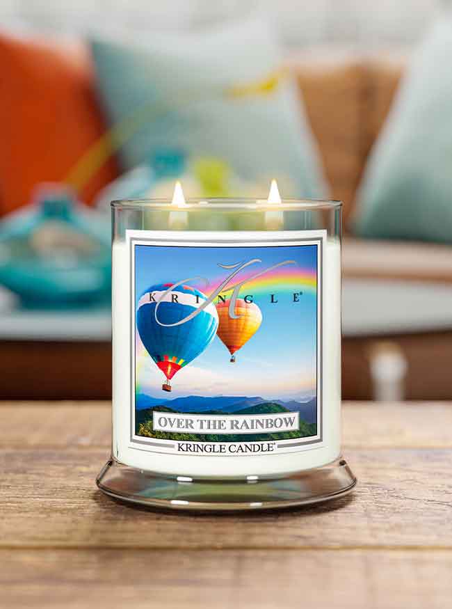A lit Kringle Candle labeled “Over the Rainbow” sits on a wooden surface. The candle jar features an image of two colorful hot air balloons floating above mountains with a vibrant rainbow arching across the sky. In the background, a cozy, softly blurred living room with teal, orange, and white pillows enhances the cheerful and uplifting atmosphere.