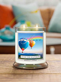 A lit Kringle Candle labeled “Over the Rainbow” sits on a wooden surface. The candle jar features an image of two colorful hot air balloons floating above mountains with a vibrant rainbow arching across the sky. In the background, a cozy, softly blurred living room with teal, orange, and white pillows enhances the cheerful and uplifting atmosphere.