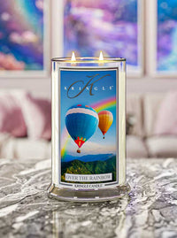 A lit Kringle Candle labeled “Over the Rainbow” sits on a polished marble surface. The candle jar features a vibrant label showing two colorful hot air balloons floating above lush green mountains with a bright rainbow in the background. Behind the candle is a softly blurred living room with pastel decor and abstract art, adding a dreamy, uplifting ambiance.