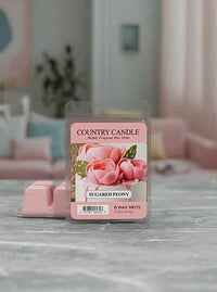 Pink wax melts labeled "Sugared Peony" by Country Candle, featuring a floral design, on a marble countertop in a cozy room.