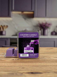 Purple wax melts package labeled "Country Candle, Grape Soda" on a wooden table with a blurred kitchen background.