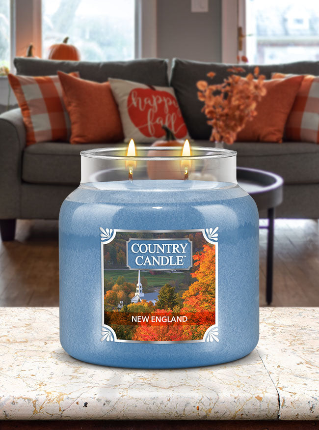 Blue jar candle labeled "Country Candle, New England" with autumn decor in a cozy living room.