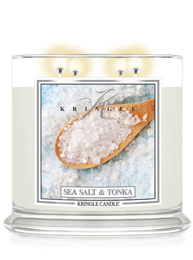 A lit Kringle Candle labeled “Sea Salt & Tonka” in a clear jar with four flames. The label features a close-up image of coarse sea salt in a wooden spoon resting on a bed of white and light blue salt crystals. The design evokes a fresh, clean, and coastal vibe against a plain white background.