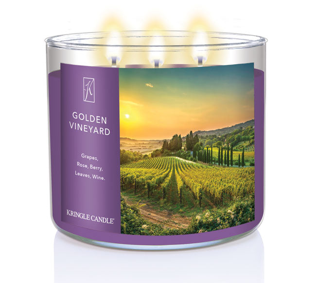 A three-wick lit candle in a clear glass jar with a scenic vineyard-themed label. The candle’s wax is deep purple, and all three wicks are burning.