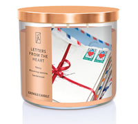 A three-wick candle in a clear glass jar with a bronze lid and a sentimental-themed label. The label features a bundle of vintage love letters tied with a red ribbon, with USA heart stamps on top. The candle’s wax is a soft peach shade, and the wicks are unlit. The scent notes listed are peony, blooming jasmine, and sandalwood.