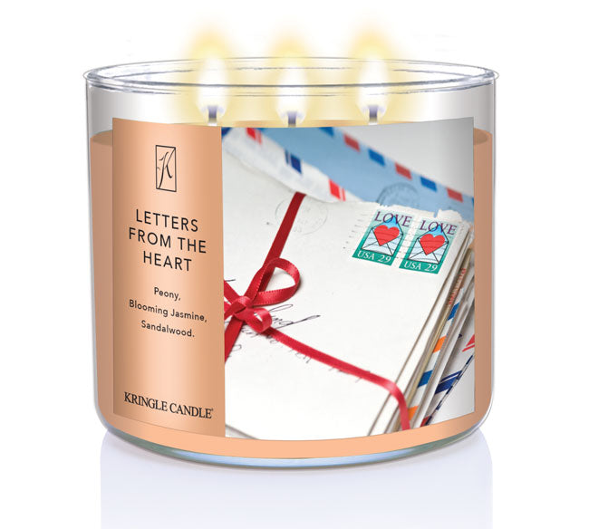 A three-wick lit candle in a clear glass jar with a sentimental-themed label. The candle’s wax is a soft peach shade, and all three wicks are burning.