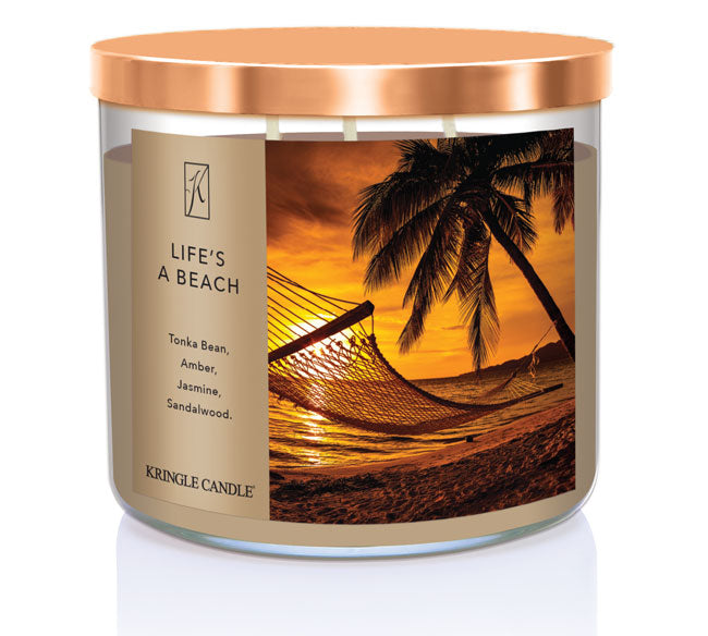 A three-wick candle in a clear glass jar with a bronze lid and a tropical sunset-themed label. The label features a hammock tied between palm trees on a golden beach at sunset, with warm orange hues in the sky. The candle’s wax is sandy beige, and the wicks are unlit. The scent notes listed are tonka bean, amber, jasmine, and sandalwood.