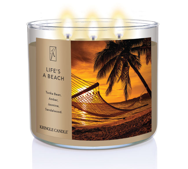 A three-wick lit candle in a clear glass jar with a tropical sunset-themed label. The candle’s wax is sandy beige, and all three wicks are burning.