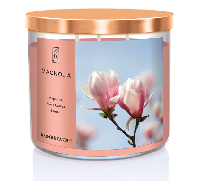 A three-wick candle in a clear glass jar with a bronze lid and a soft floral-themed label. The label features a close-up of pink magnolia blossoms against a bright blue sky. The candle’s wax is a warm peach shade, and the wicks are unlit. The scent notes listed are magnolia, fresh leaves, and lemon.