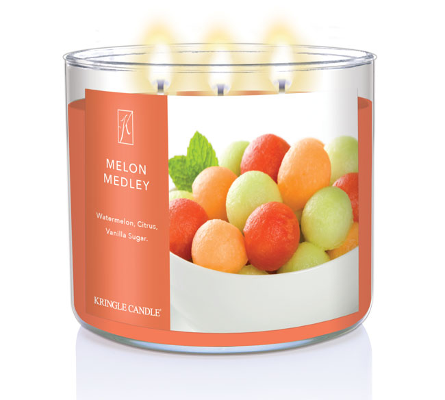 A three-wick lit candle in a clear glass jar with a fresh fruit-themed label. The candle’s wax is warm orange, and all three wicks are burning.