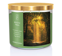 A three-wick candle in a clear glass jar with a gold lid and a forest-inspired label. The label features a golden-lit forest path with autumn leaves and sun rays filtering through the trees. The candle’s wax is an earthy green, and the wicks are unlit. The scent notes listed are fresh berries, apple, autumn leaves, caramel & spice sugar, and woods.