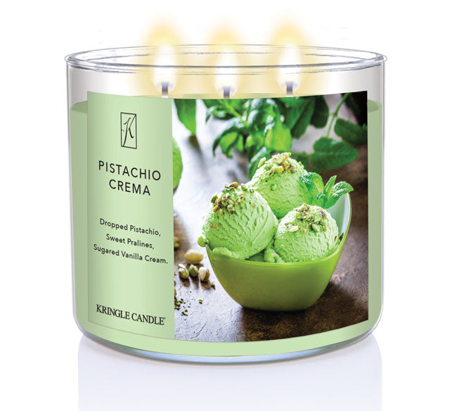 A three-wick lit candle in a clear glass jar with a creamy dessert-themed label. The candle’s wax is soft green, and all three wicks are burning.