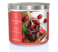 A three-wick candle in a clear glass jar with a black lid and a sweet jam-themed label. The label features a glass jar of strawberry jam with fresh strawberries scattered around. The candle’s wax is deep red, and the wicks are unlit. The scent notes listed are strawberries, tart lemon, and vanilla buttercream.