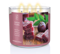 A three-wick lit candle in a clear glass jar with a festive holiday-themed label. The candle’s wax is deep purple, and all three wicks are burning.