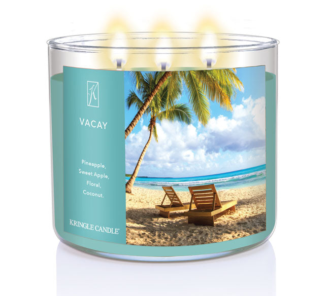 A three-wick lit candle in a clear glass jar with a tropical vacation-themed label. The candle’s wax is sunny yellow, and all three wicks are burning.
