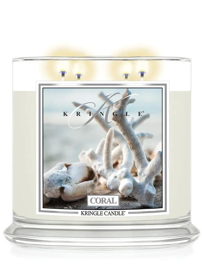 Lit Kringle Candle in Coral scent with glowing flames and label showing white coral on a beach background.