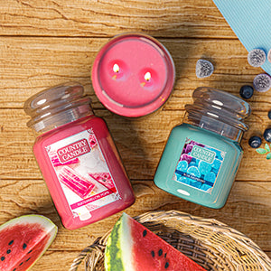 This image for the Country Candle Fruit Collection has two large candle jars, one candles' two wicks are lit. The labels show a strawberry mint tart and blueberry ice cream. The candles set on a kitchen counter with rustic wood cabinets. 