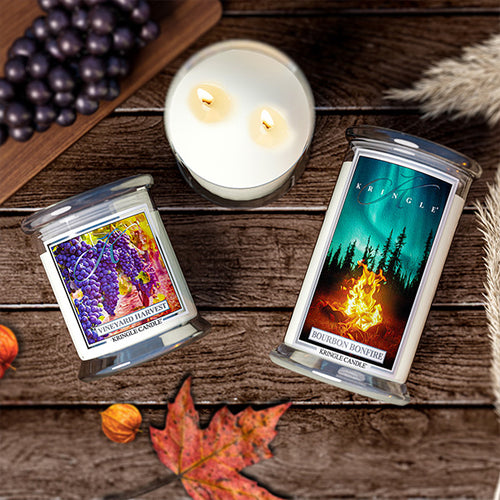 This image represents Kringle Candle Autumn collection with two large fall candles with pumpkins and red and orange leaves on trees and ground. The candles are set in a living room with a couch and wall art.