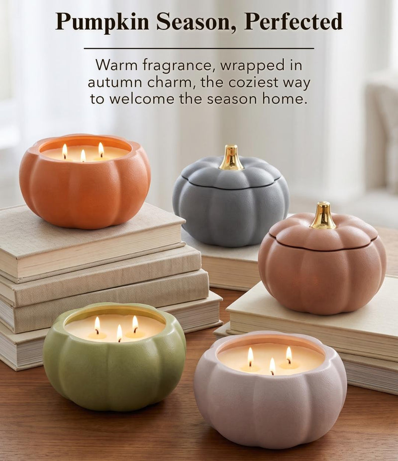 Four pumpkin-shaped candle holders in orange, gray, sage green, and blush pink, arranged on stacked books on a wooden surface. Two are open with lit candles and two have lids on.