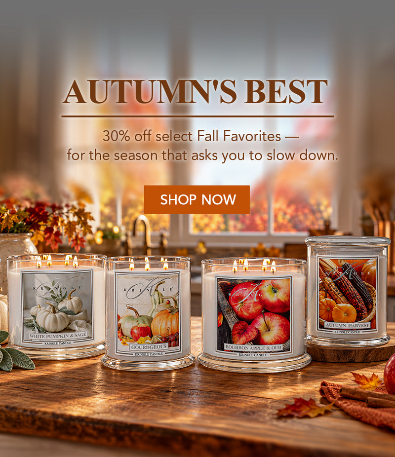 Four lit scented candles in clear glass jars are arranged on a wooden table in a warm autumn-themed setting. The labels show fall illustrations including pumpkins, apples, and corn.