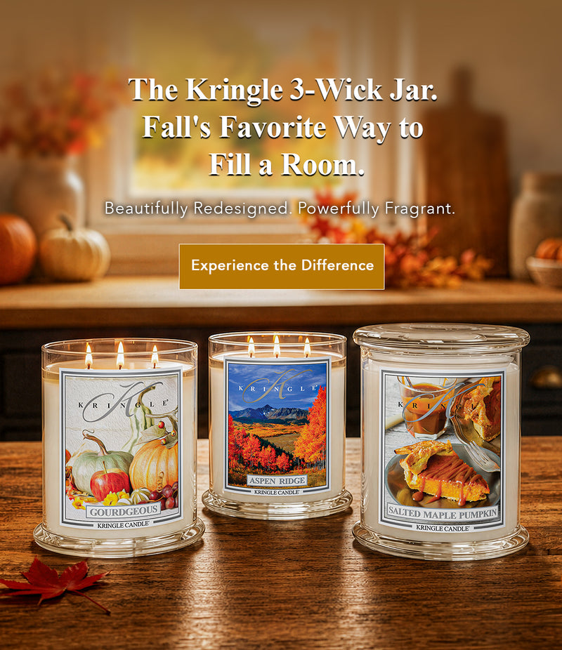 Three lit 3-wick candles in clear glass jars displayed on a wooden table in a warm autumn-themed room setting. The labels feature fall illustrations: 'Ligh Horizons' with pumpkins and apples, 'Alpine Ridge' with mountain and autumn scenery, and 'Salted Maple Pumpkin' with pumpkin pie imagery.