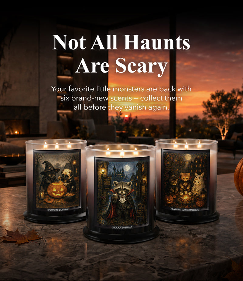 Three lit Halloween-themed scented candles in large glass jars sit on a stone table in a cozy indoor setting at sunset. The candles feature illustrated labels with pumpkins, a raccoon in a cape, and cats under a full moon.